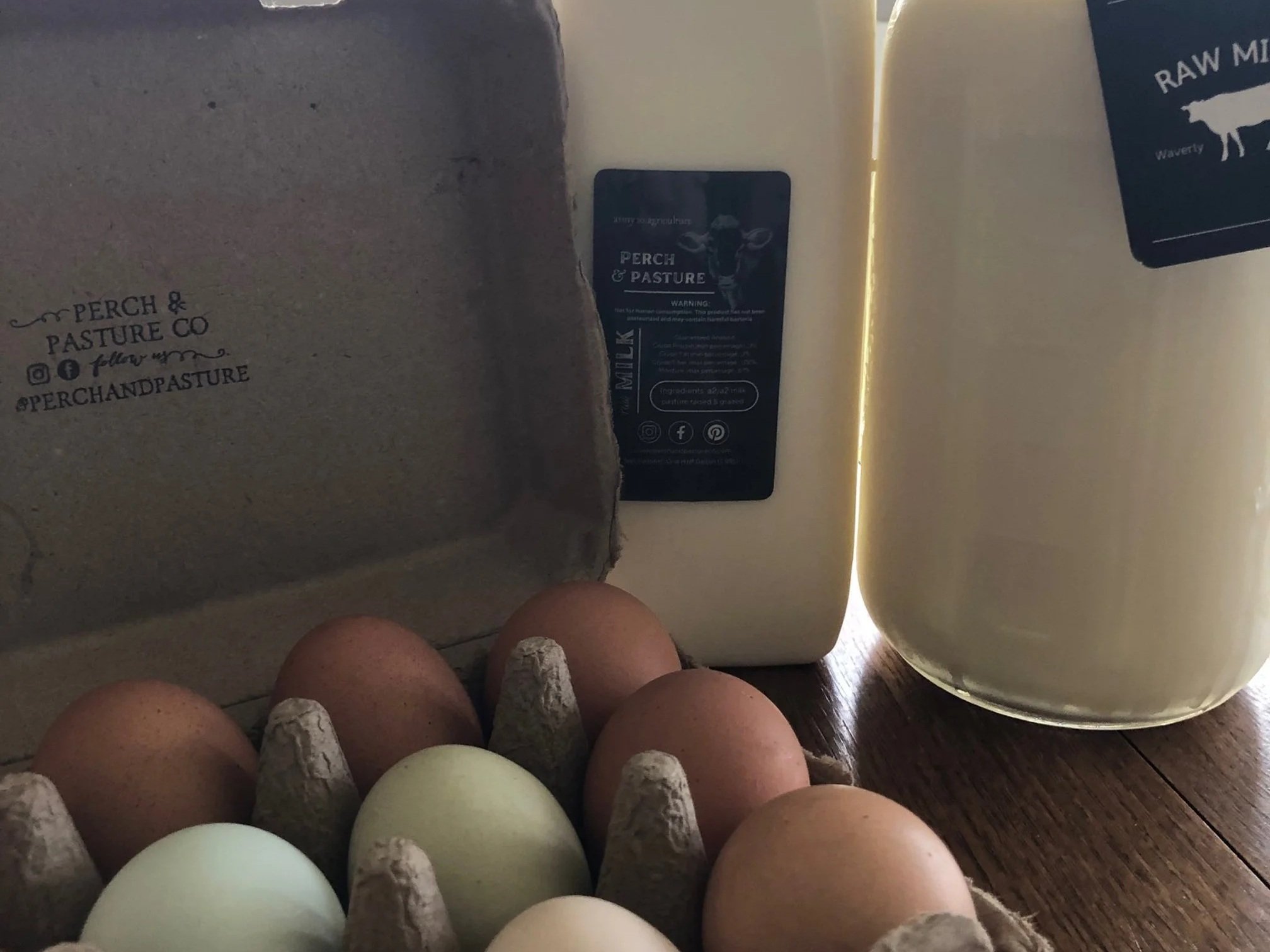 Farm-fresh eggs in a carton next to bottles of raw milk from Perch & Pasture Co, showcasing a variety of egg colors and the company branding.