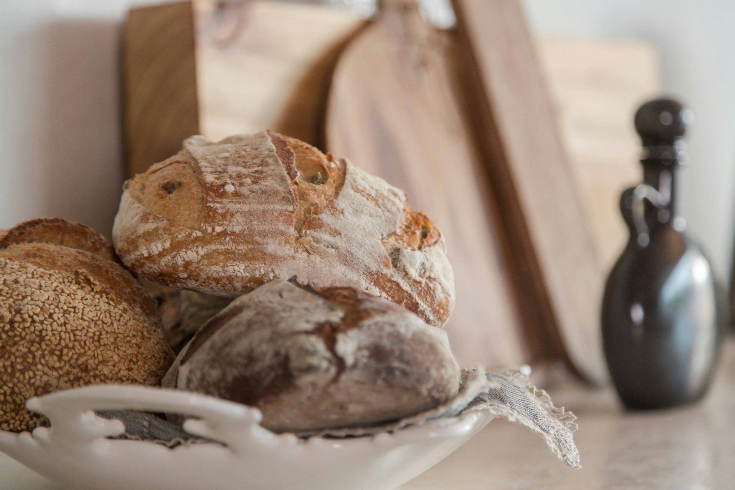 Rustic bread varieties displayed in a white ceramic bowl, featuring sesame, crusty, and whole grain loaves, with wooden cutting boards and a dark ceramic oil dispenser in the background. Perfect for a cozy kitchen setting or bakery aesthetic.