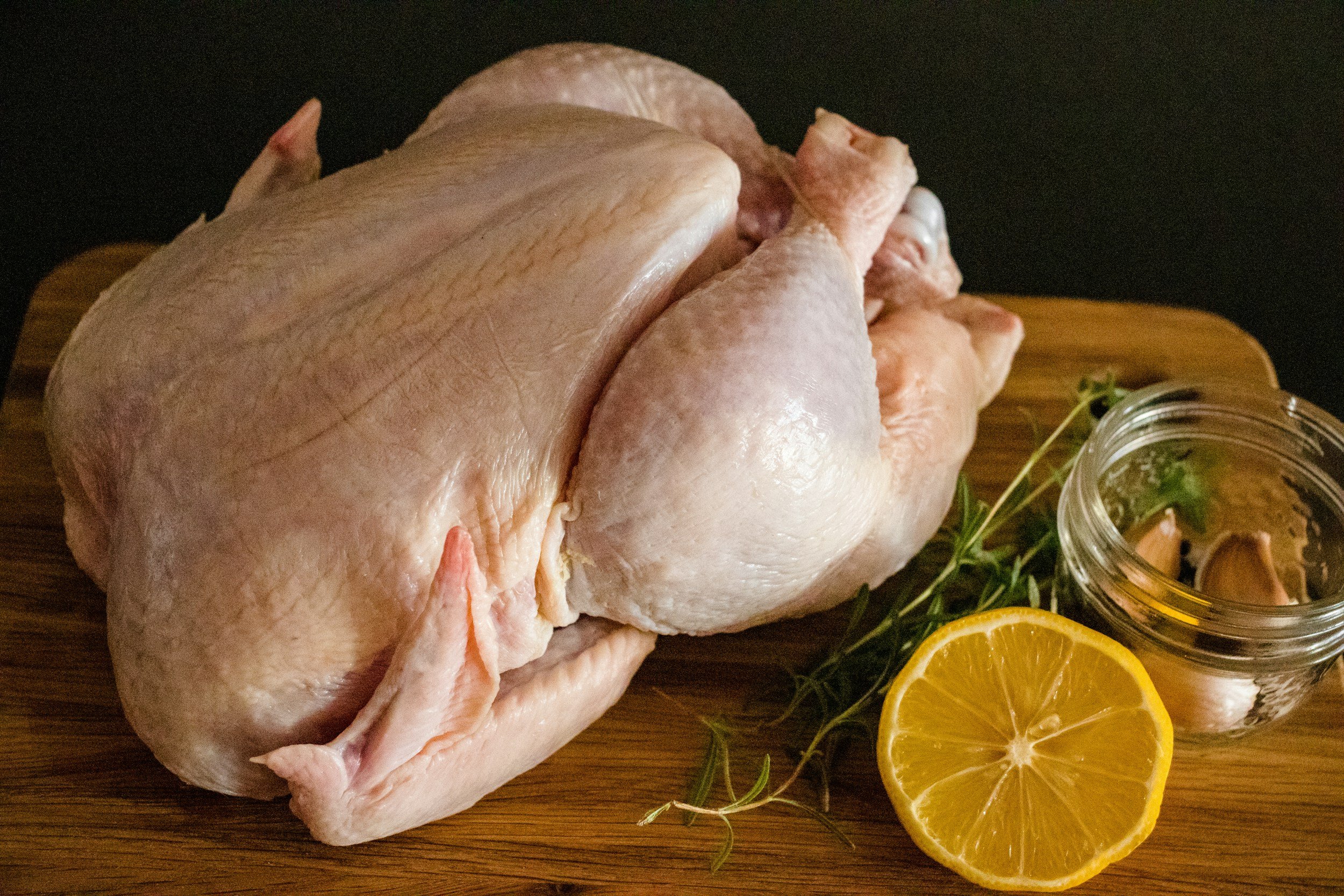 Raw whole chicken on a wooden cutting board, accompanied by fresh lemon slices, garlic cloves, and herbs, ideal for cooking or meal preparation.