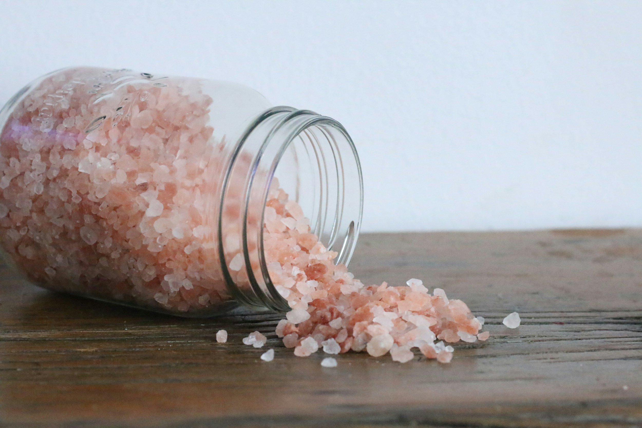 Close-up of a glass jar spilling pink Himalayan salt onto a wooden surface, showcasing the coarse texture and natural color of the salt crystals against a neutral background.