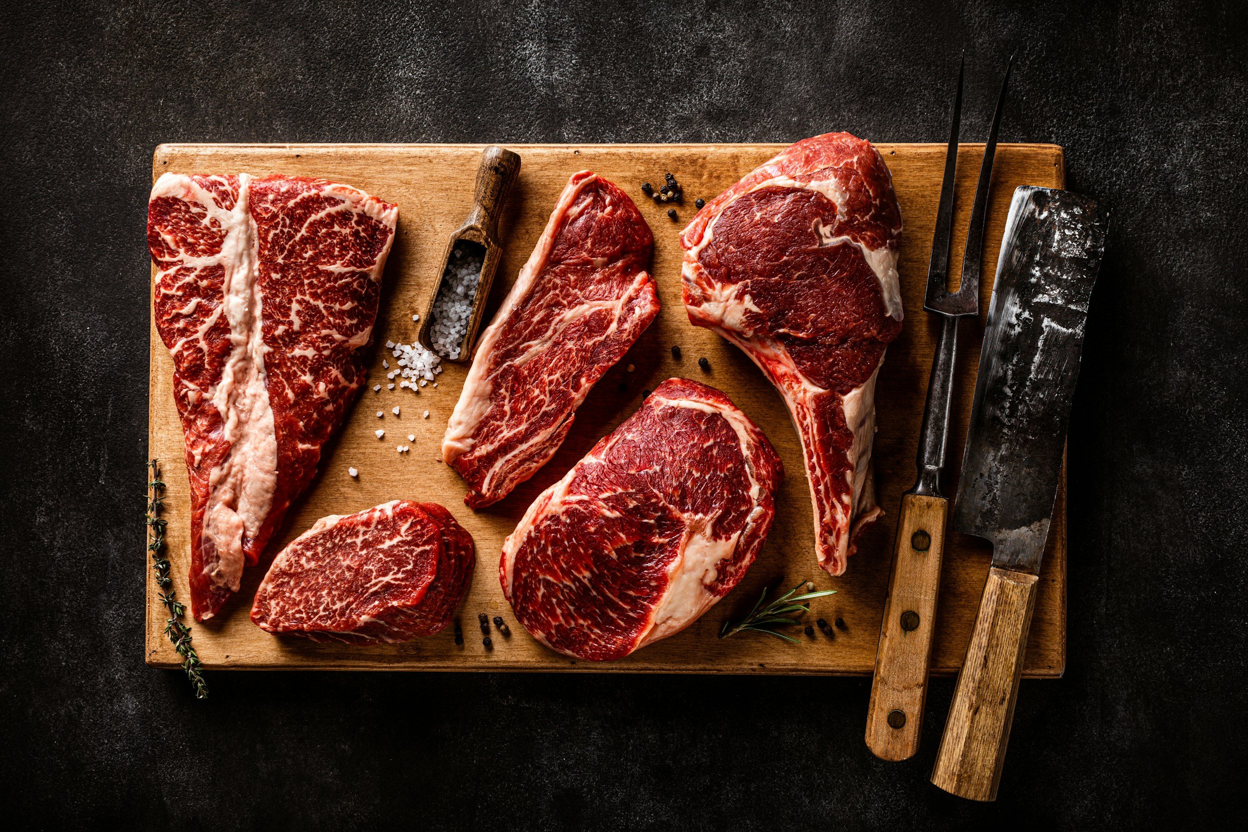 Assorted cuts of high-quality beef including ribeye, sirloin, and T-bone, displayed on a wooden cutting board with a butcher's knife and seasoning, ideal for gourmet cooking and grilling.